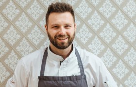 Philipp Essl in chef's clothes in front of a patterned wallpaper.