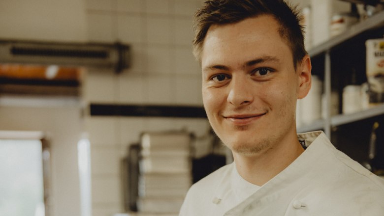 A young man in a white chef's jacket smiles into the camera in a kitchen.