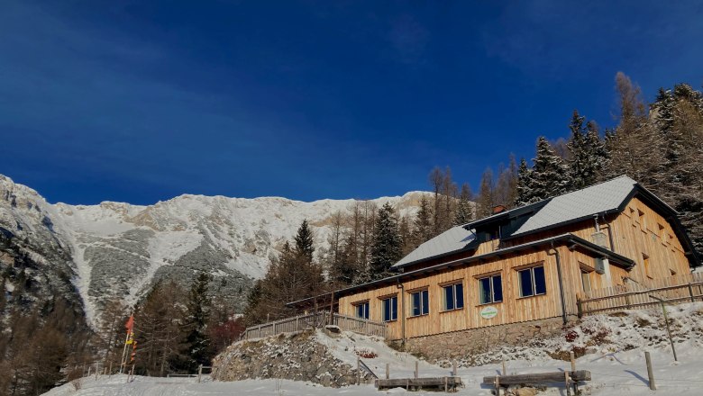 Waxriegelhaus, © Martin Tod - Waxriegelhaus A wooden house in the snow against a mountain backdrop under a clear blue sky.
