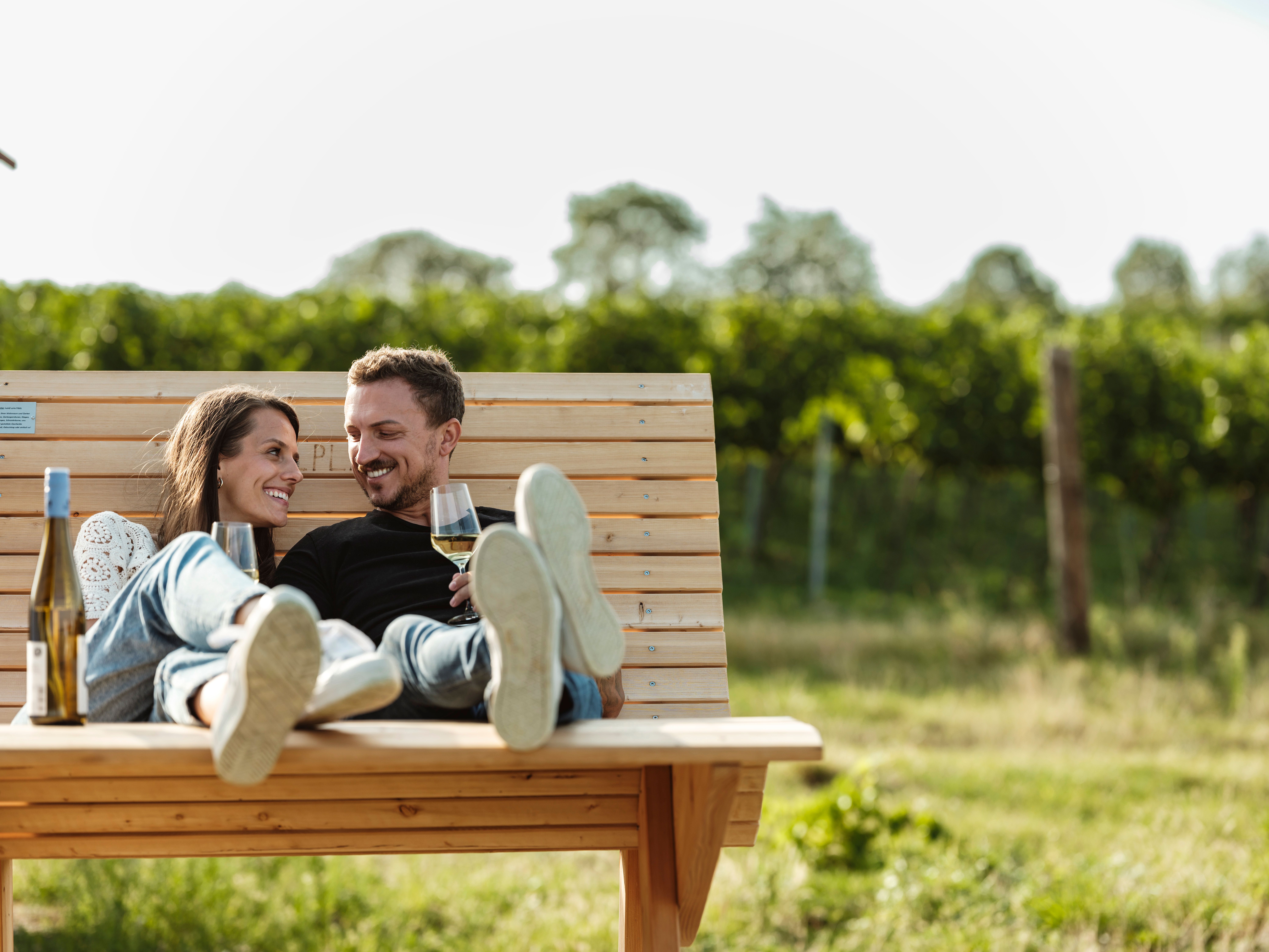 A couple relaxes on a wooden bench outside with glasses of wine in their hands.
