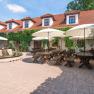 A cozy courtyard of a winery with wooden tables, benches and parasols, surrounded by a building with red tiled roofs and green vegetation.