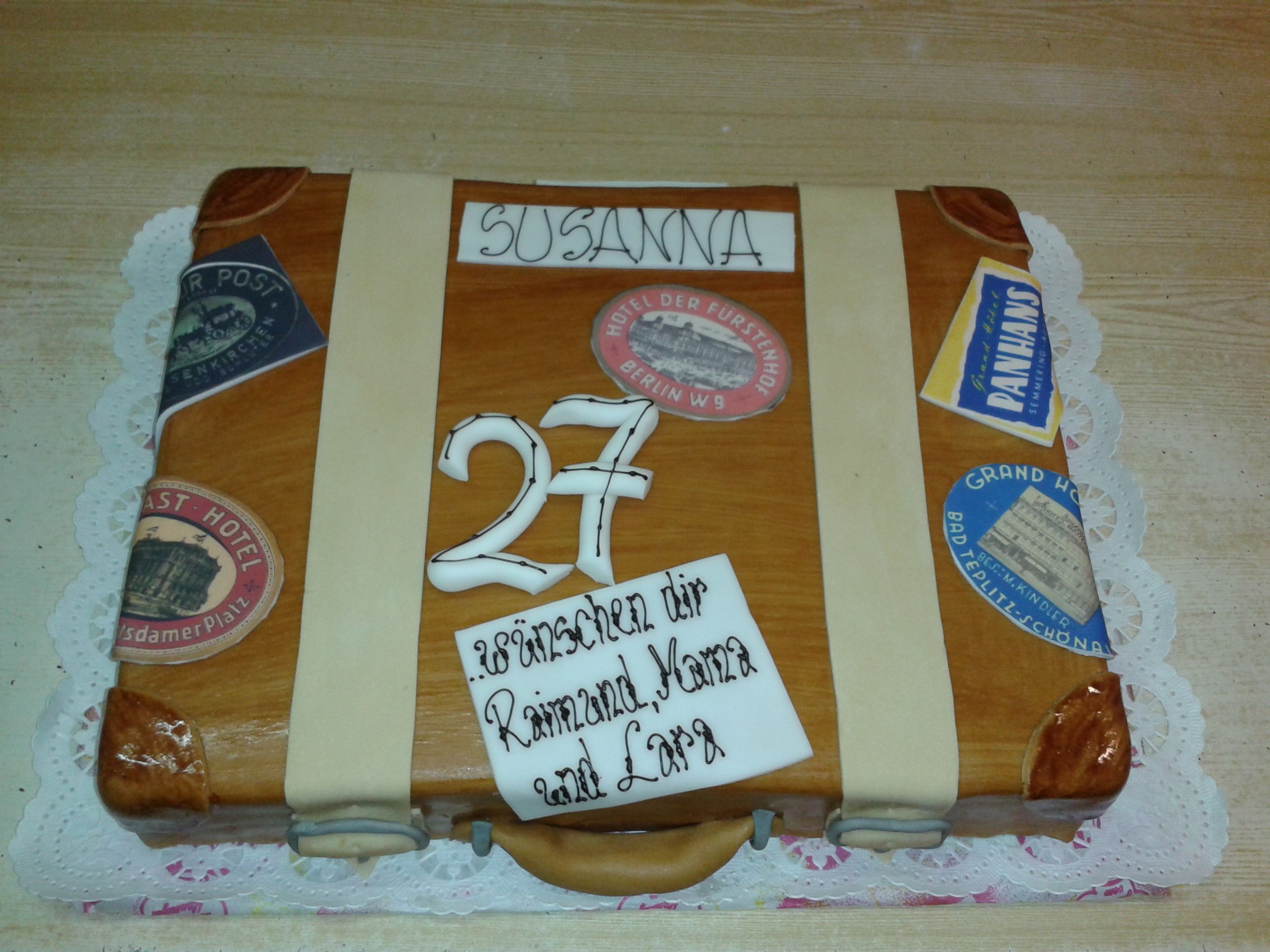 A cake in the shape of a suitcase with stickers and the number 27.
