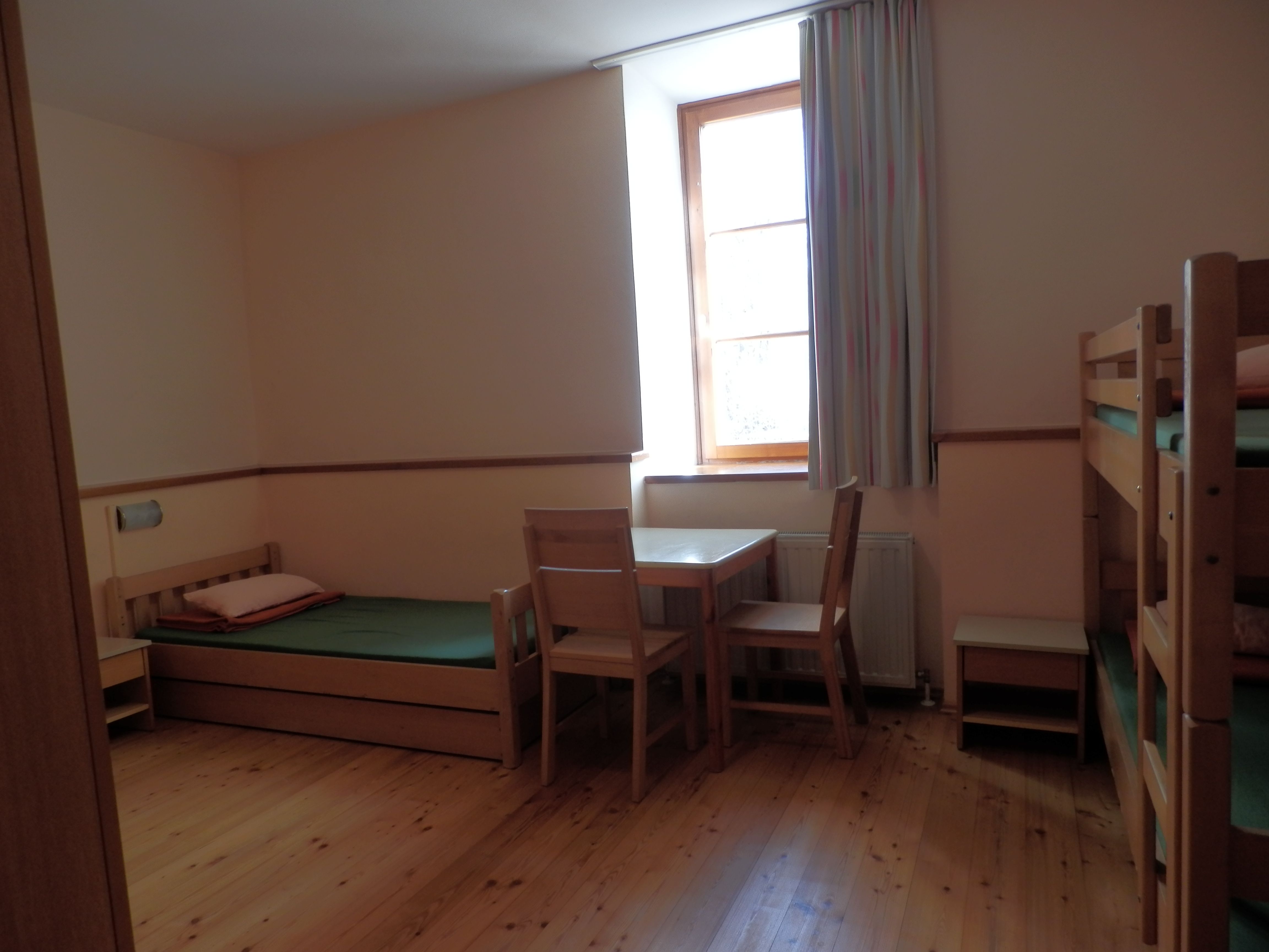 A simply furnished guest room with wooden floor, single bed, bunk bed, table and chairs, and a window with curtain.