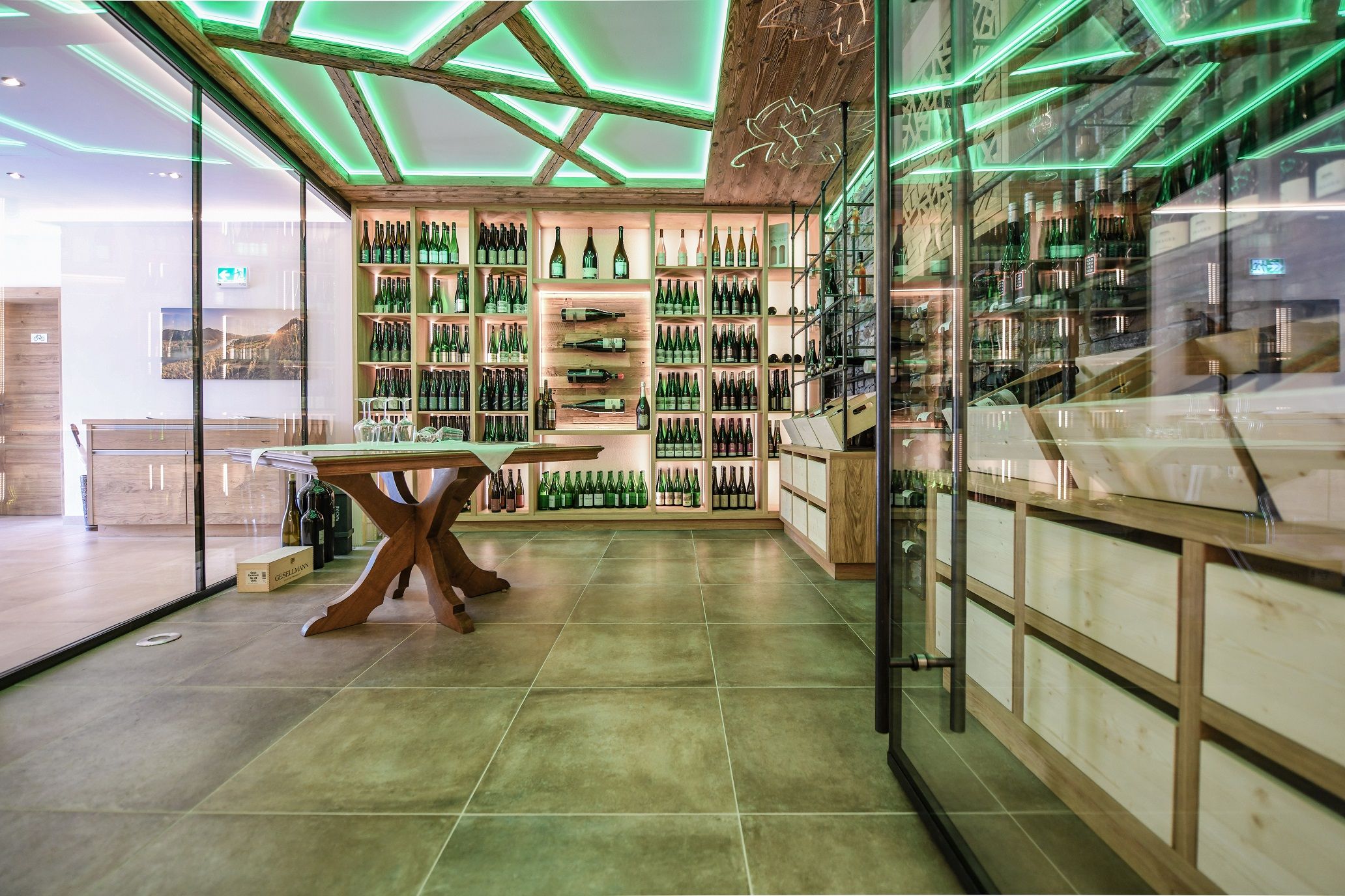 Modern wine presentation room with glass walls, wooden shelves full of wine bottles and a green illuminated ceiling.