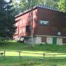A red building with wooden cladding, surrounded by trees and a meadow, with a sign "Naturfreundehaus Ortsgruppe Ternitz".