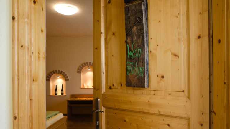 Open wooden door with sign 'Wein-Frühling', view into a cozy room with wine bottles in niches.