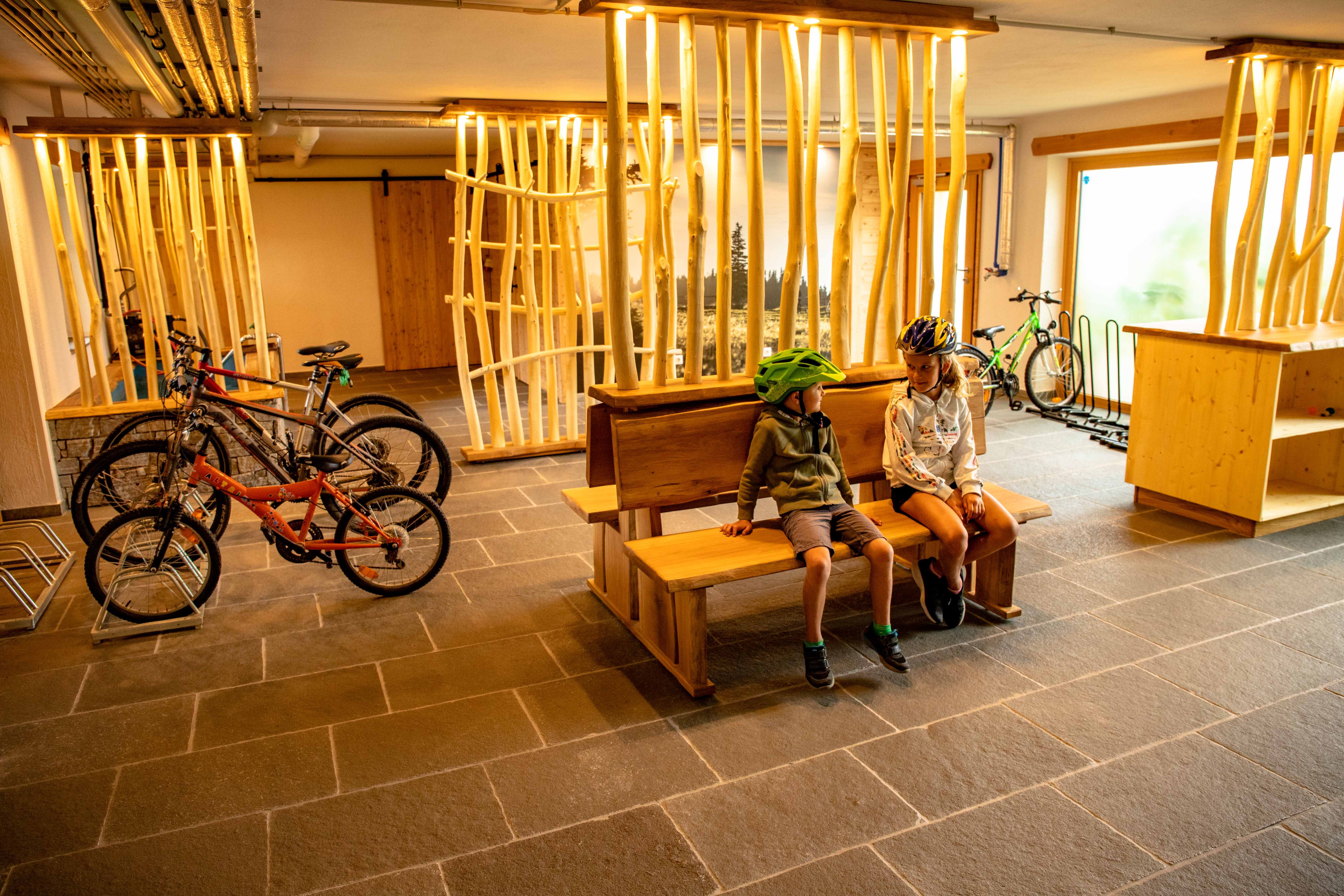 Two children with bicycle helmets sit on a wooden bench in a room with bicycles and wooden decorations.