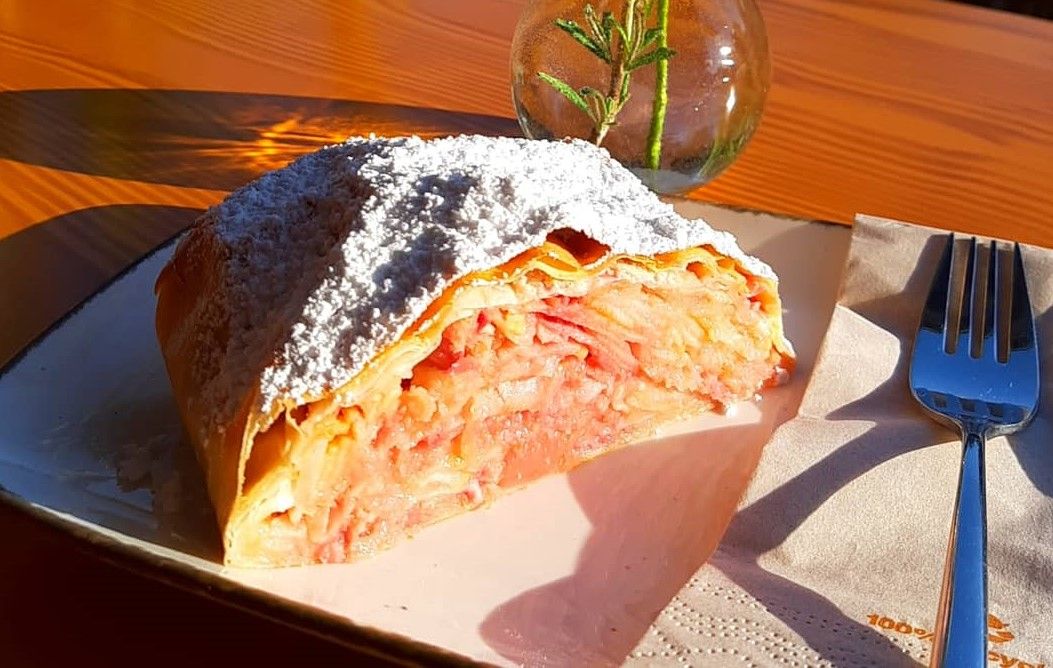 A piece of apple strudel with powdered sugar on a plate, next to it a fork and a vase of rosemary.