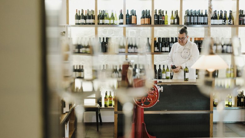 Man in a wine shop, surrounded by bottles of wine, looking at a bottle.