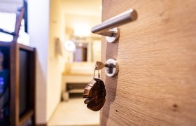 Close-up of a wooden door with a key in the lock carrying a wooden pendant. A blurred room can be seen in the background.