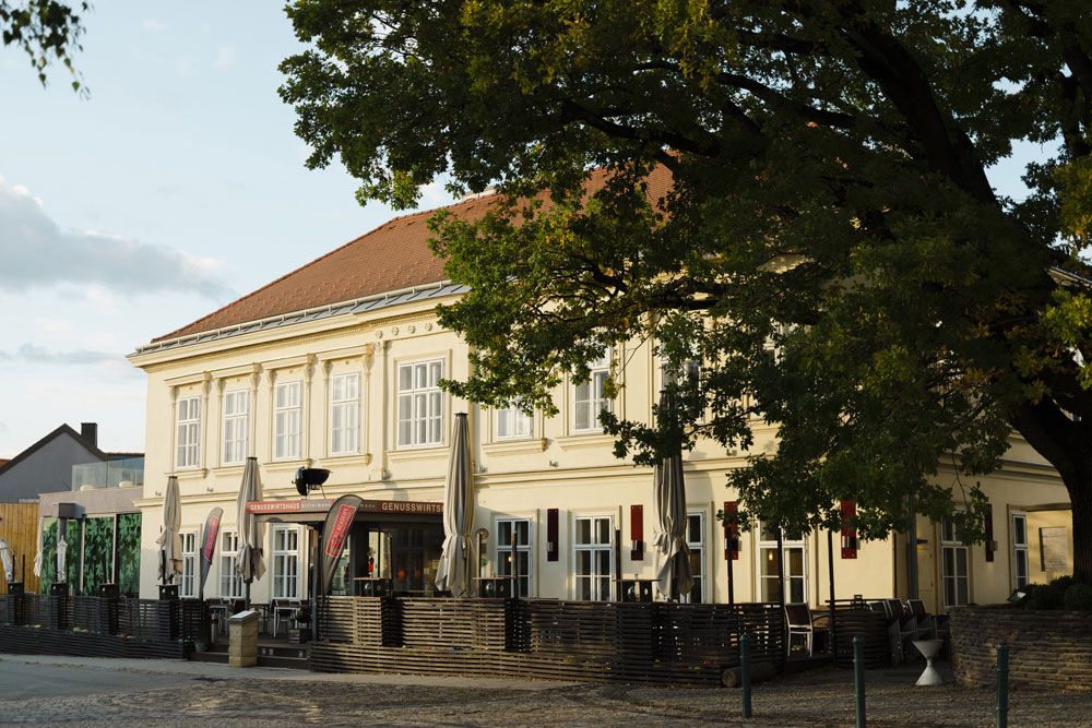 A large, two-storey building with a terrace and parasols, surrounded by trees.