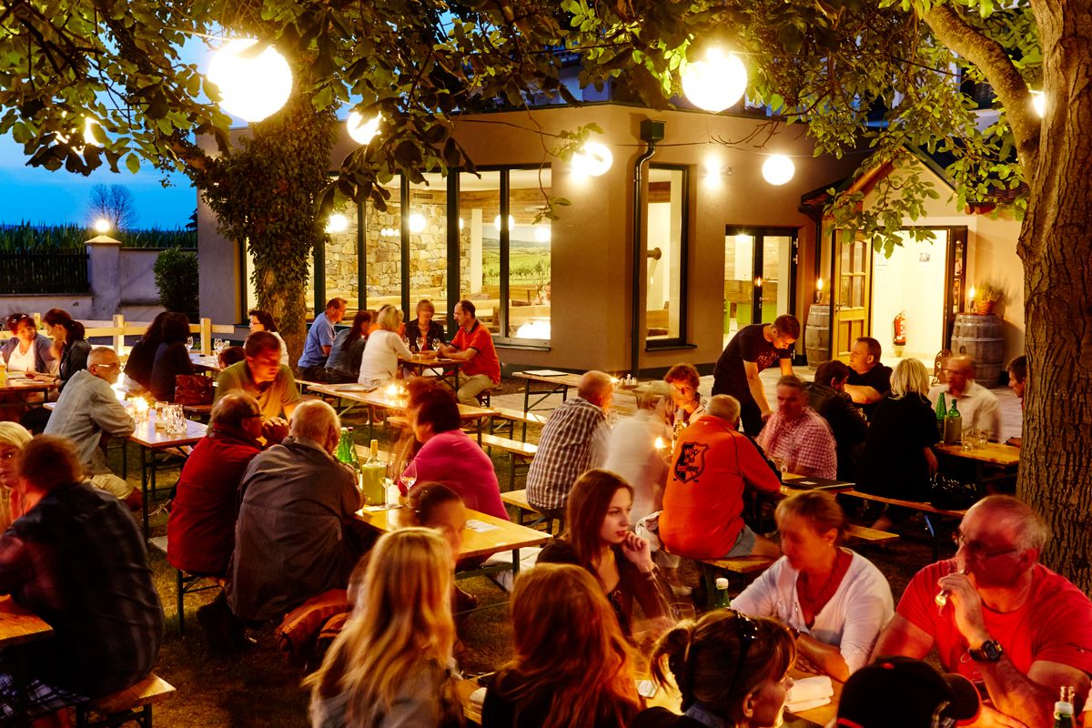People sit at outdoor tables in the evening, surrounded by trees and lights.