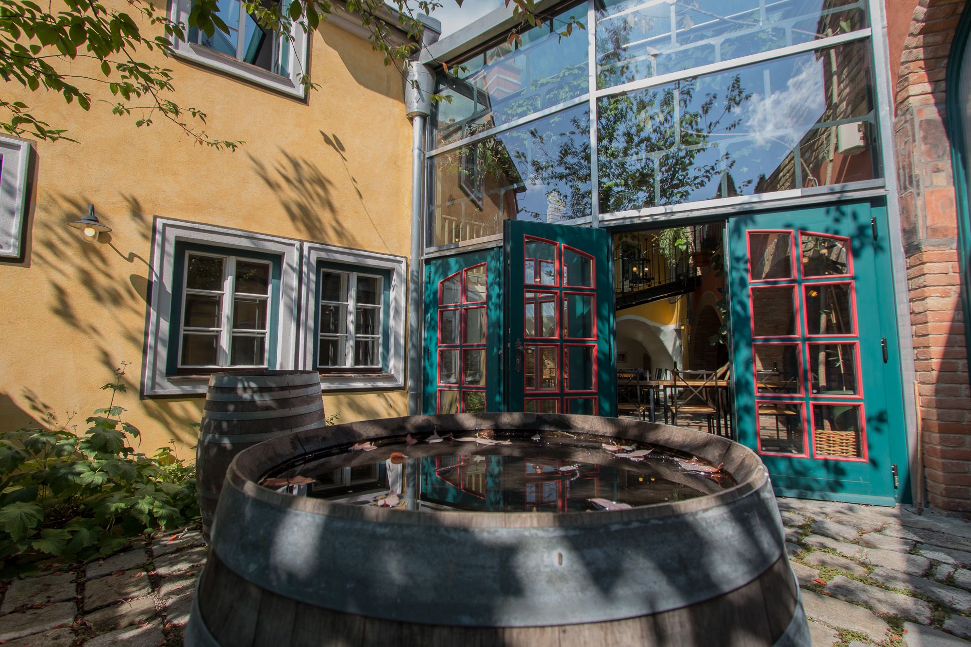 An inner courtyard with a yellow façade, green doors and a wooden barrel of water. Shadows of trees fall on the wall.