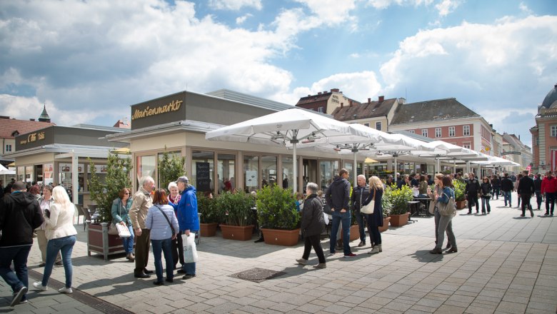 People on a busy market square with cafés and stores under parasols.