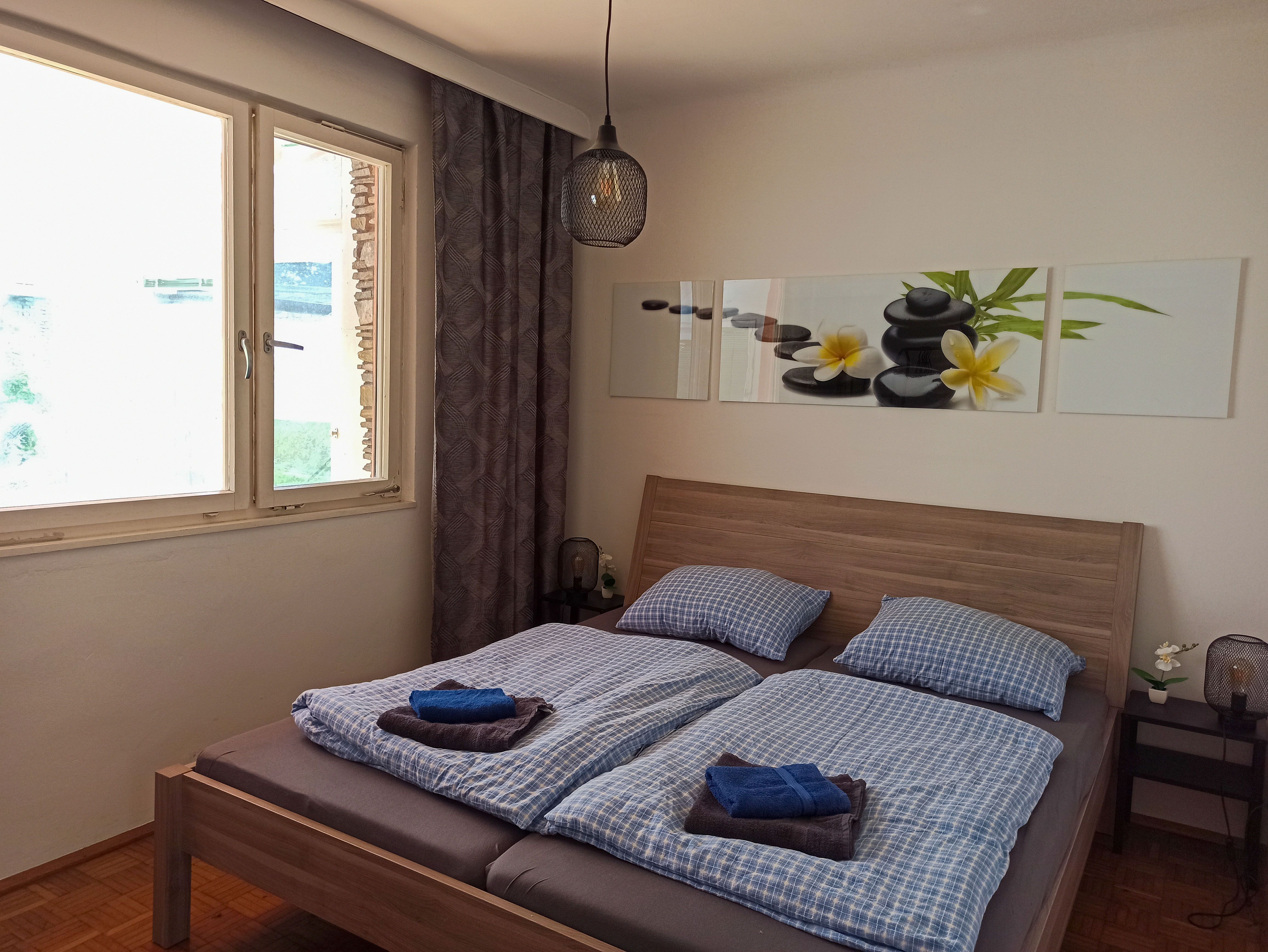 Bedroom with double bed, blue bed linen, picture with stones and flowers on the wall, window and bedside lamp.