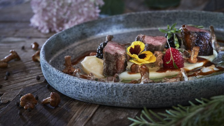 A plate of venison fillet, chanterelles, berries and edible flowers on a wooden table.