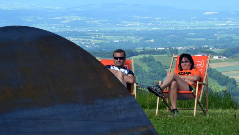 Two people relax in deckchairs on a lawn with a view of a vast landscape.