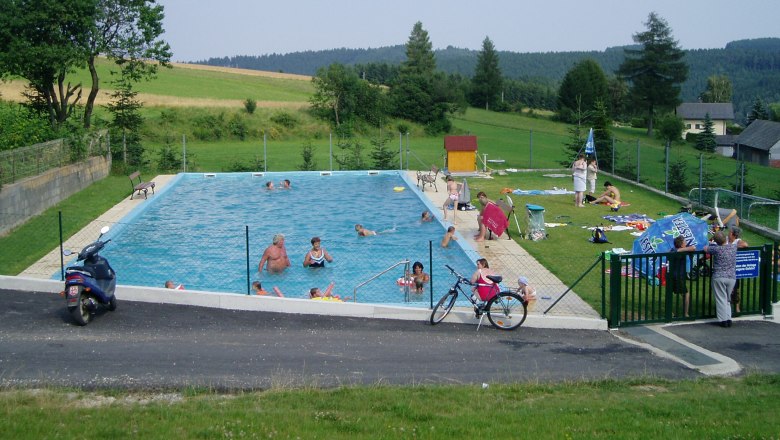 An outdoor pool with several people, surrounded by green landscape and trees.