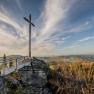 A summit cross on a rock with a viewing platform, surrounded by forest and a wide view over the landscape under a blue sky.