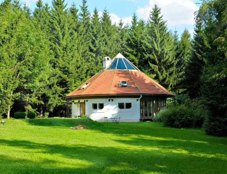 A round, white house with a pointed roof in the middle of a green forest.