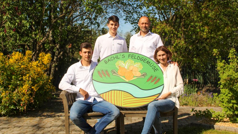 Four people pose with a sign for 'Bachl's Feldgem&uuml;se' in a garden.