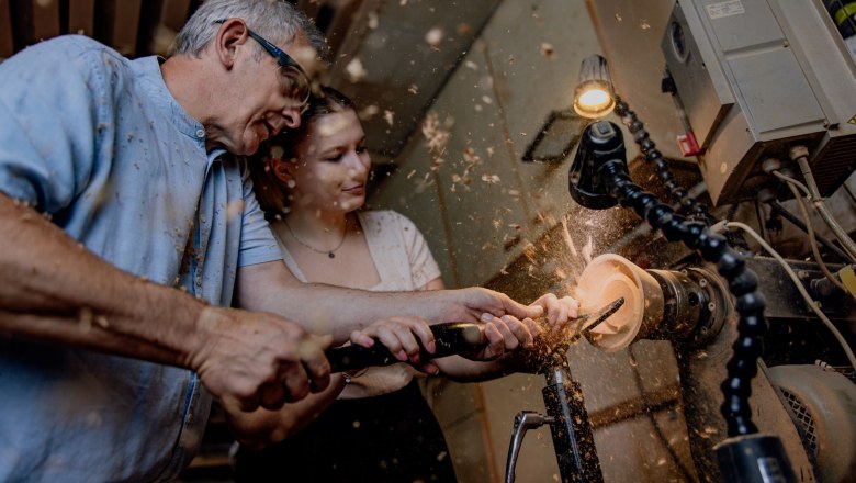 Two people work together at a lathe to turn a piece of wood. Wood shavings fly in the air.