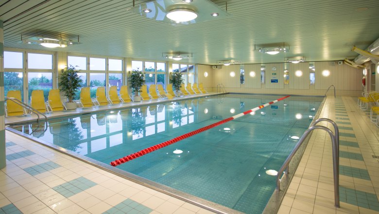 Interior view of an empty indoor pool with yellow deckchairs and plants.