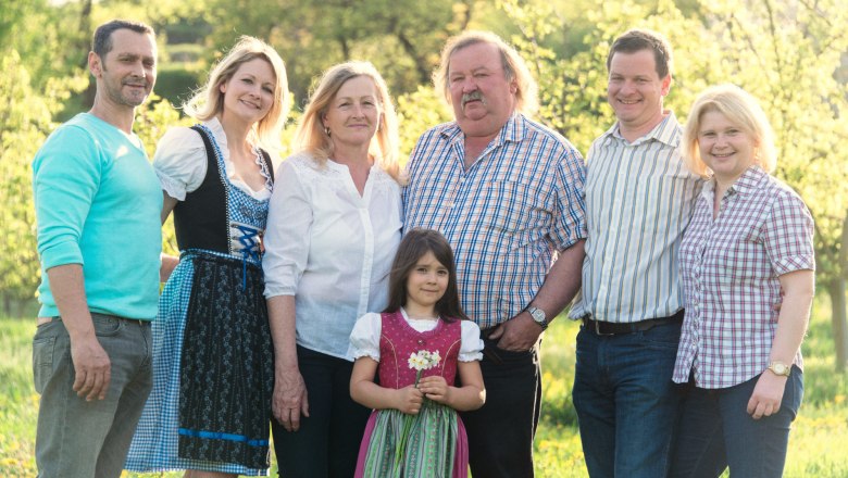 A smiling family stands in a sunny garden, some wearing traditional clothing.
