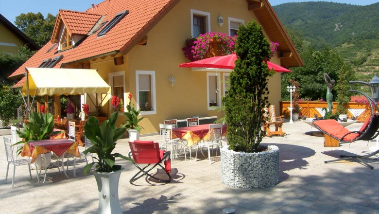 Guest house "Zur schönen Aussicht", © Elisabeth Wegscheider Guest house with terrace, tables, chairs and parasols, surrounded by plants and mountains in the background.