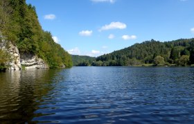 A calm lake surrounded by wooded hills under a blue sky with few clouds.