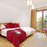 Bright bedroom with double bed, red pillows and blanket, wooden floor and large window with curtains.