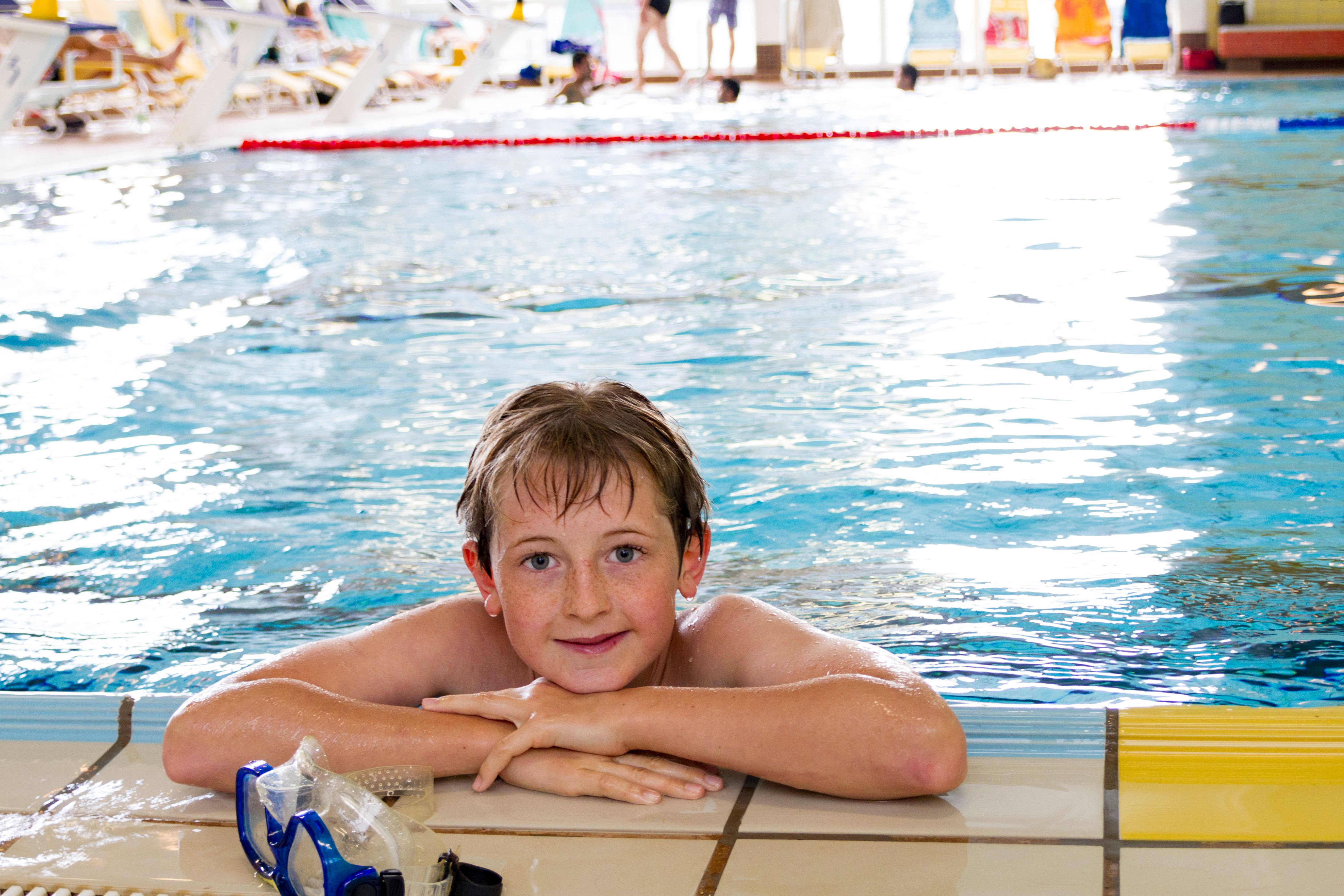 A boy is leaning against the edge of an indoor pool, smiling and wearing swimming goggles.