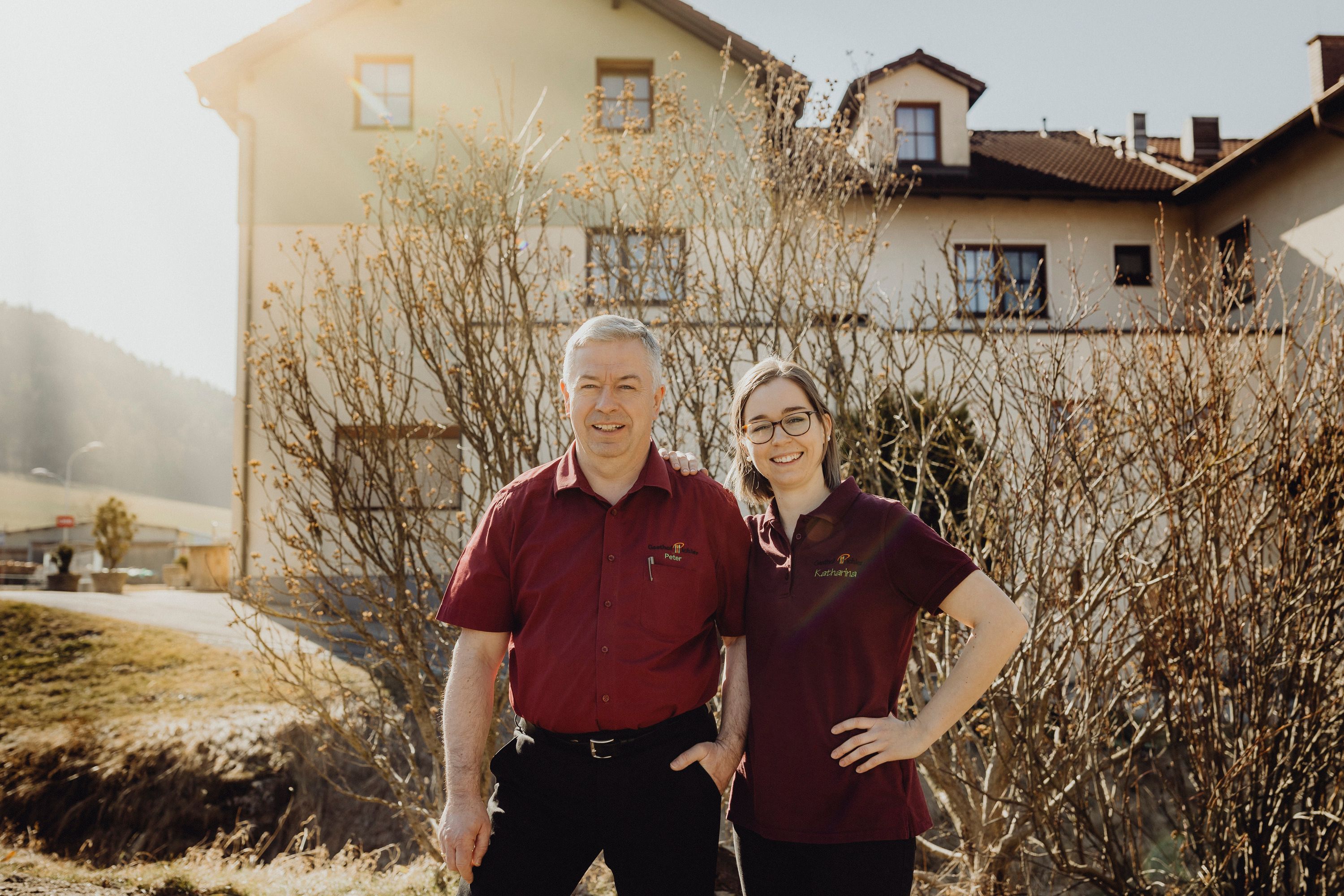 Two people in red shirts stand smiling in front of a building.