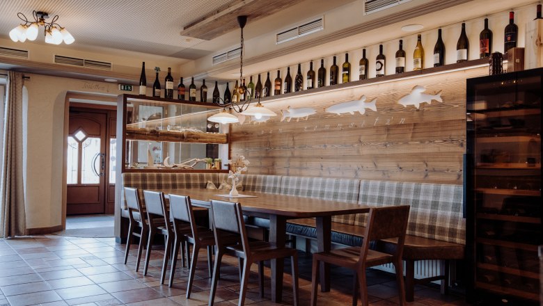 Cozy dining area with wooden table, chairs and bench, decorated with wine bottles and fish motifs on the wall.