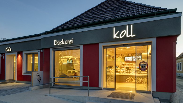 Exterior view of the bakery in the evening with illuminated sign 'Bäckerei Koll' and 'Cafe'.
