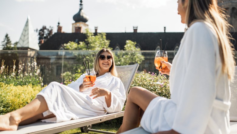 Two women in bathrobes relax on deckchairs with drinks in front of a castle.
