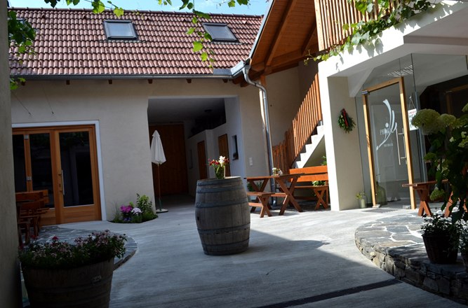 A cozy courtyard with wooden furniture, plants and a wine barrel. A building with red roof tiles and a wooden staircase in the background.