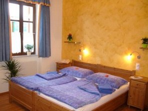 A cozy double room with wooden furniture, blue bed linen and yellow wall. Plants and windows with blue curtains are visible.