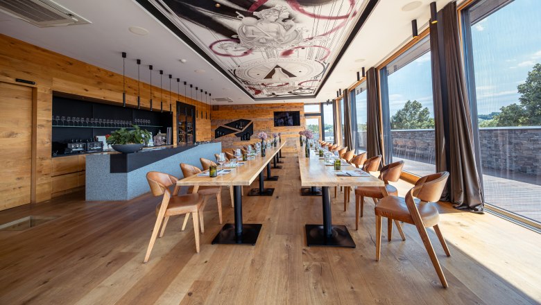 Interior view of a modern restaurant with long wooden tables, leather armchairs and large windows.