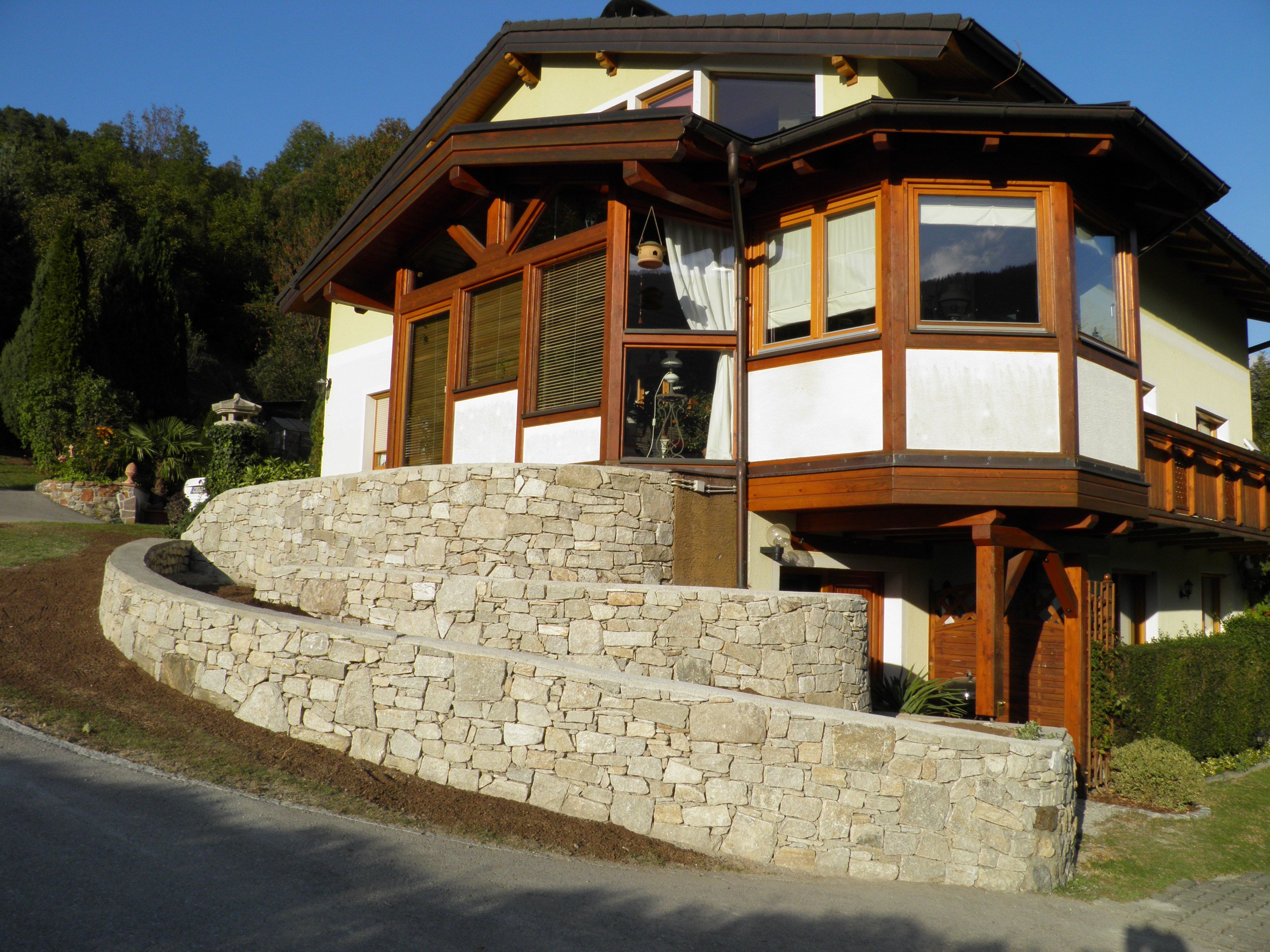 House with stone wall and wooden cladding, surrounded by trees.