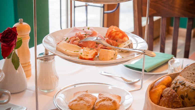 A breakfast table with an etagere full of cold cuts, jams and bread rolls, next to a glass of orange juice.