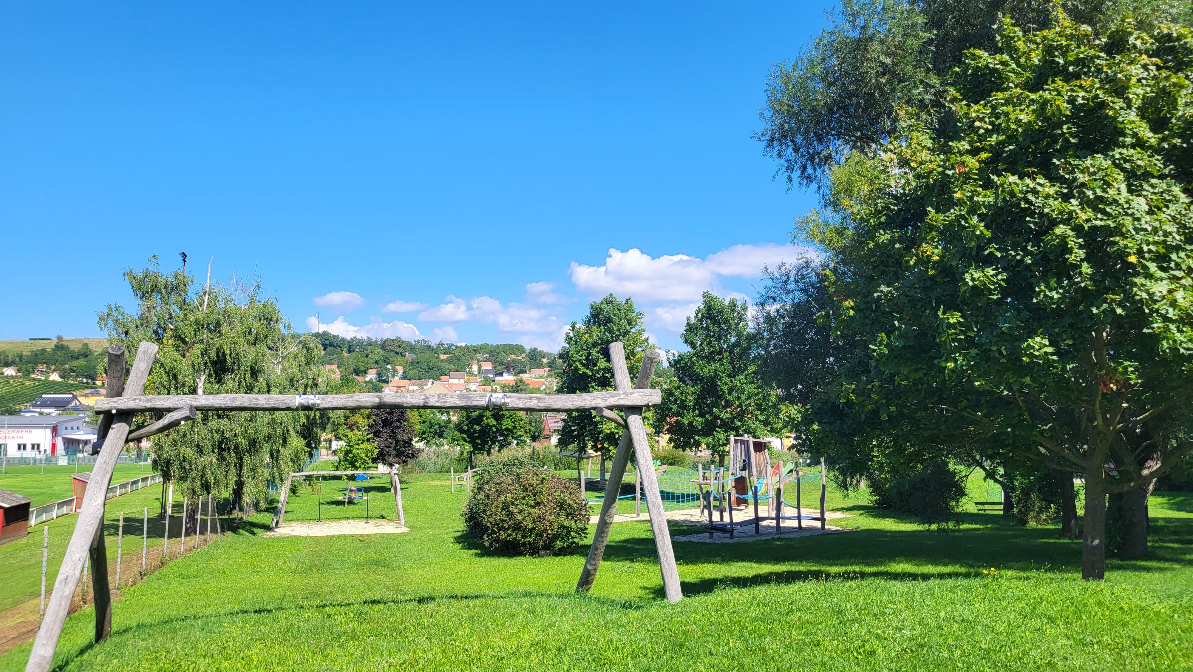 A playground with swings and a climbing frame on a green meadow, surrounded by trees and a clear blue sky.