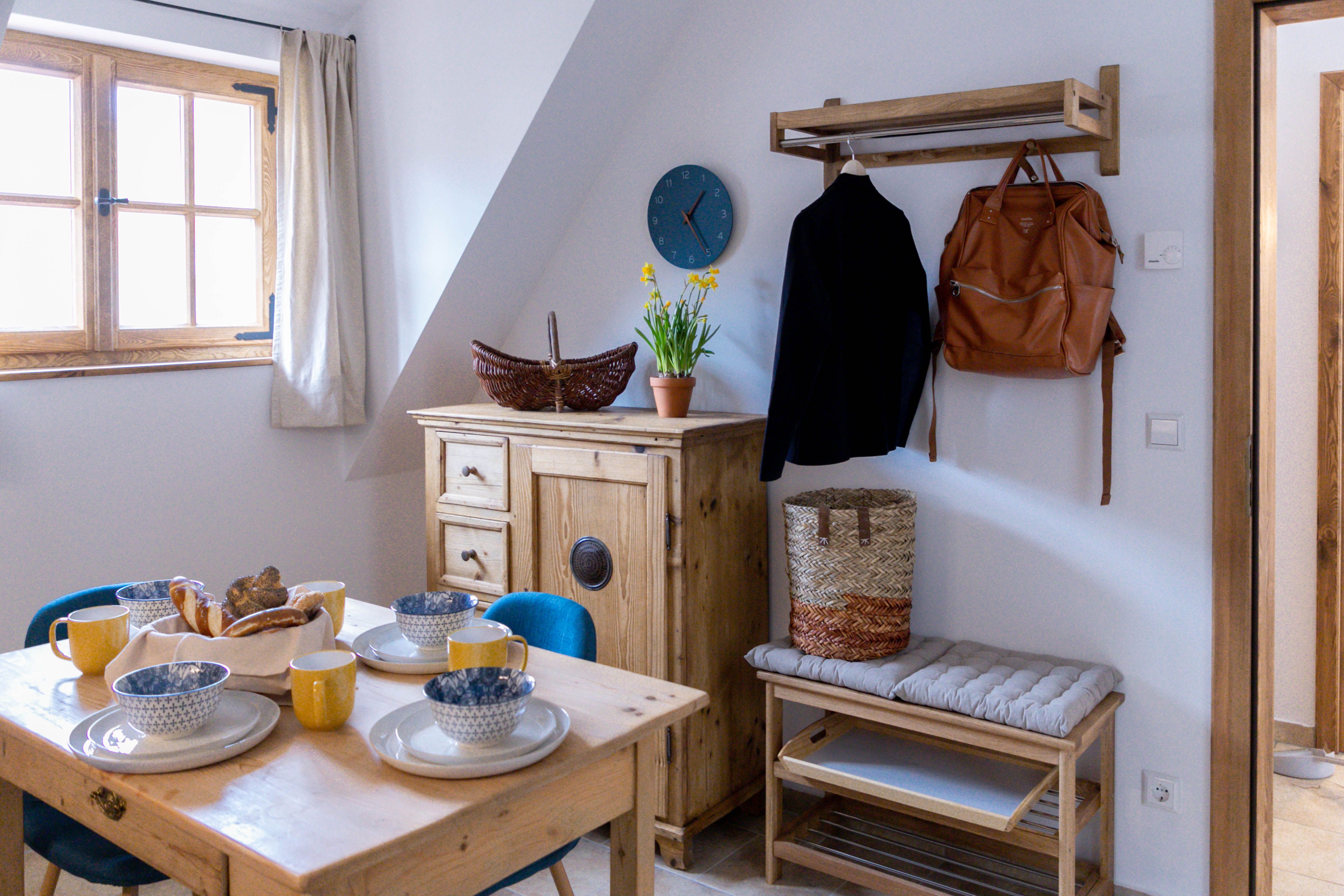 Cozy breakfast corner with wooden table, dishes, yellow cups, chest of drawers, jacket, rucksack and flowers on a cupboard.