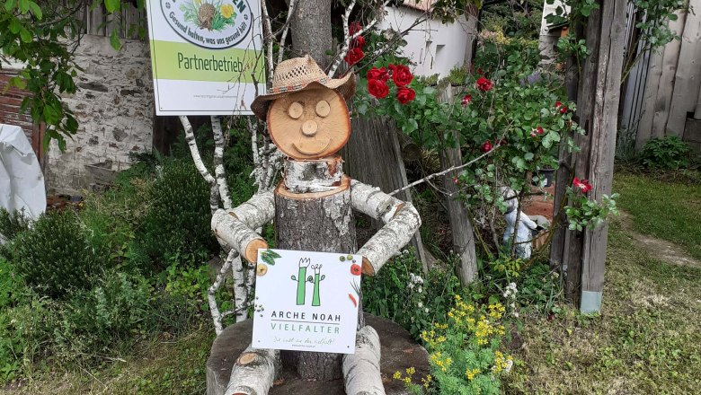 Wooden figure with straw hat sitting on tree stump, holding sign with inscription 'Arche Noah Vielfalter'. Background: Nature in the garden sign
