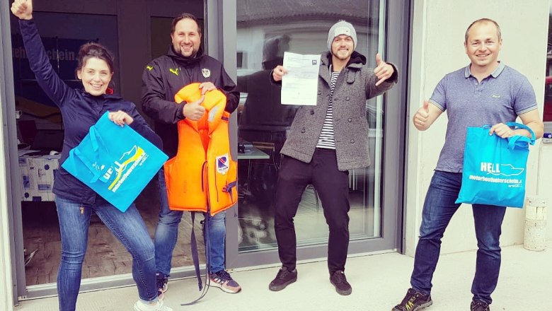 Four people are standing in front of a building, holding bags and a life jacket, smiling and giving the thumbs up.