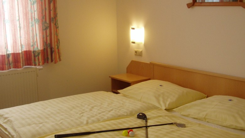 A double bed with yellow bed linen, on which lie two golf clubs and a golf ball. A window with a curtain and a picture on the wall.