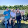 Three people are standing in front of a vineyard next to a trailer.