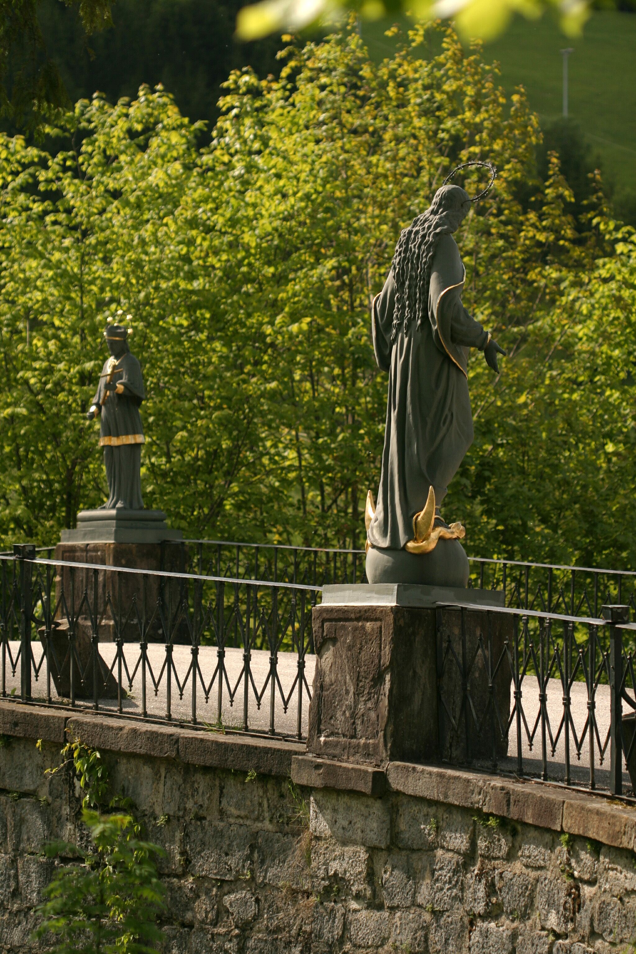 Two statues on a bridge with railings, surrounded by green trees.