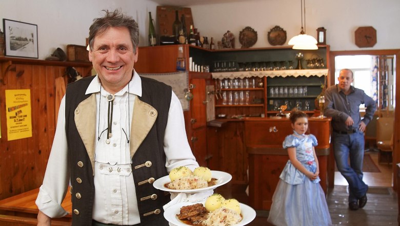 A man in traditional dress serves roast pork with dumplings in a rustic inn. In the background a man and a girl in a dress.