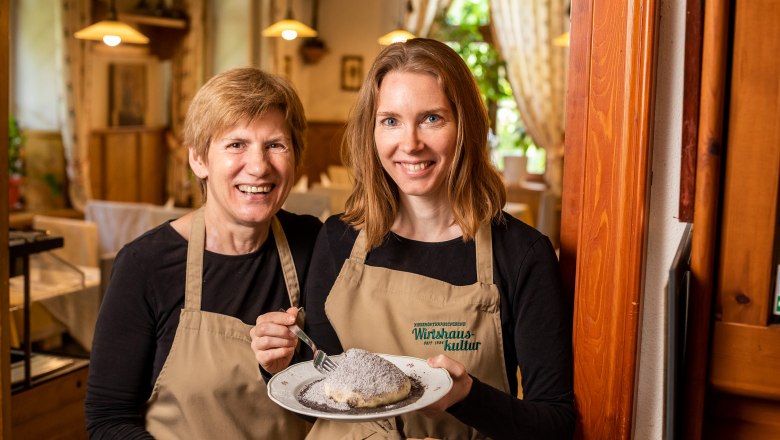 Two women in aprons hold a plate with a dessert in a cozy restaurant.